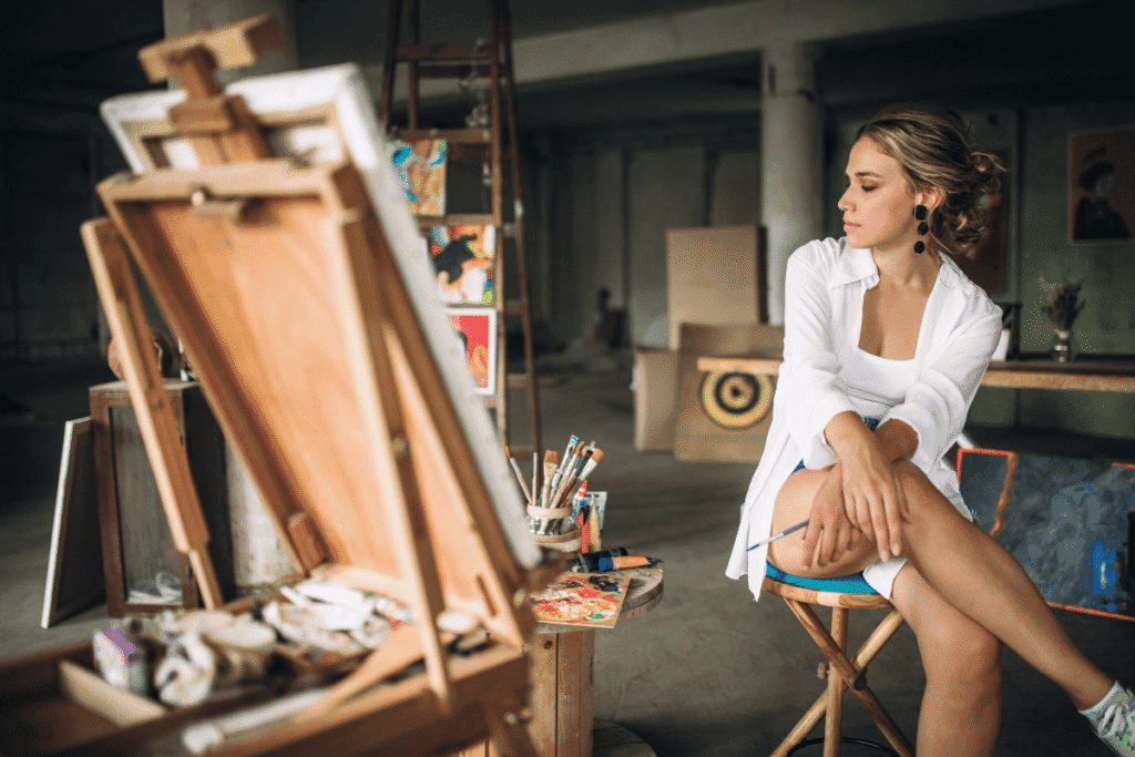 artista pensativa- com várias tintas e telas de pintura ao redor. segurando um pincel.
